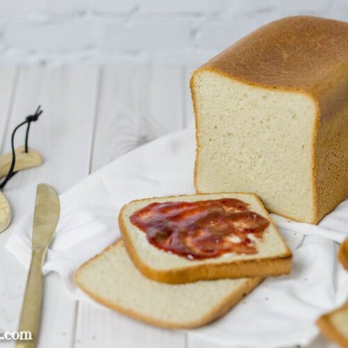 Ein Laib Toastbrot aufgeschnitten. Zwei Scheiben liegen vor dem Brot. Eine davon ist mit Butter und Marmelade bestrichen. Daneben liegt ein goldenes Messer.