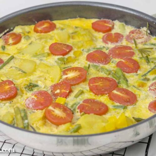Frittata mit grünem Spargel und Kartoffeln in einer Pfanne, die auf einem schwarzen Abkühlgitter steht. Davor ist ein wenig frischer Rosmarin zu sehen.