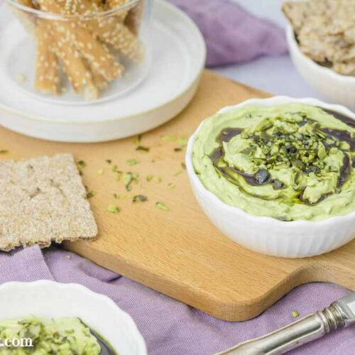Ein Schälchen mit Kürbiskernölaufstrich mit Frischkäse steht auf einem Jausenbrett. Im Hintergrund sind Grissinis und Knäckebrotstücke zu sehen. Ein violettes Tuch lässt die grüne Farbe des Dips richtig leuchten.